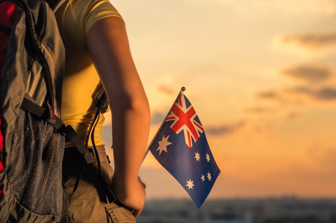 Backpacker wearing a yellow shirt and large rucksack holding a small Australian flag at sunset, representing a working holiday in Australia